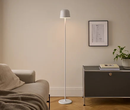Un salon moderne avec un canapé beige, un lampadaire blanc et un meuble gris foncé. La lampe allumée se tient entre le canapé et le meuble, qui supporte des livres et une plante en pot sous un tableau encadré.