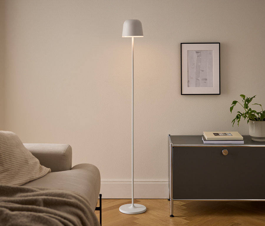Un salon moderne avec un canapé beige, un lampadaire blanc et un meuble gris foncé. La lampe allumée se tient entre le canapé et le meuble, qui supporte des livres et une plante en pot sous un tableau encadré.