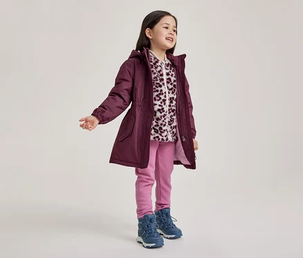 Une jeune fille se tient debout, regardant à droite et souriant, vêtue d'une parka prune à capuche, d'une polaire imprimé léopard blanc et violet, d'un pantalon rose et de bottes bleues.