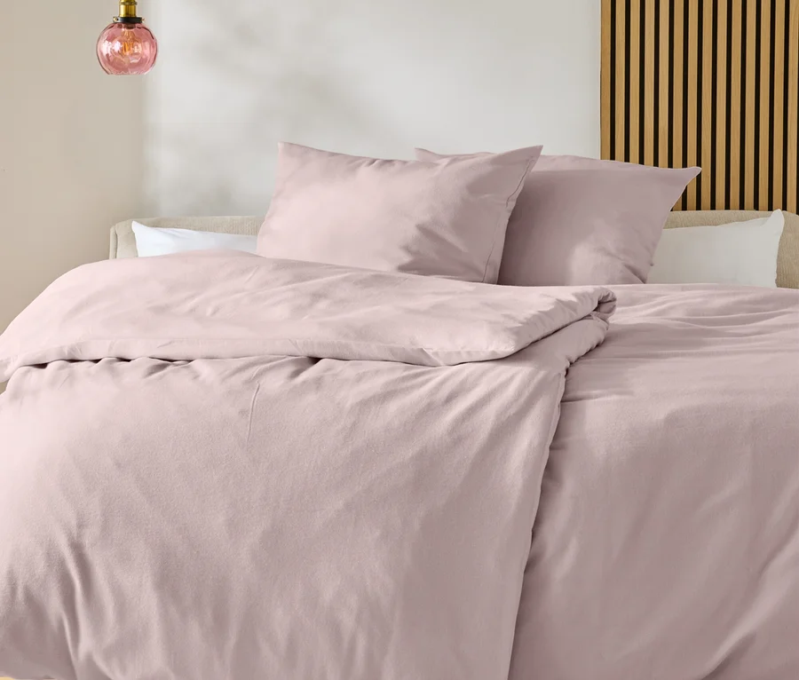Un lit avec une parure de lit rose clair et deux oreillers, devant un tapis blanc à motif noir sur un plancher en bois. Une suspension luminaire en verre rose est à gauche, un panneau mural à lattes en bois est à droite.