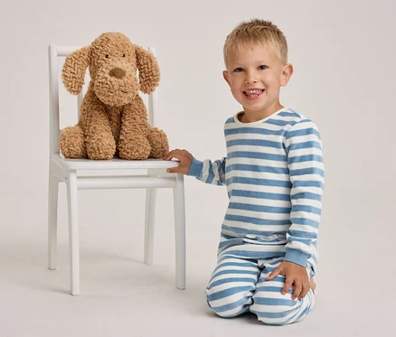 Un petit garçon souriant en pyjama rayé bleu et blanc est agenouillé à côté d'une chaise blanche sur laquelle est assis un chien en peluche marron.
