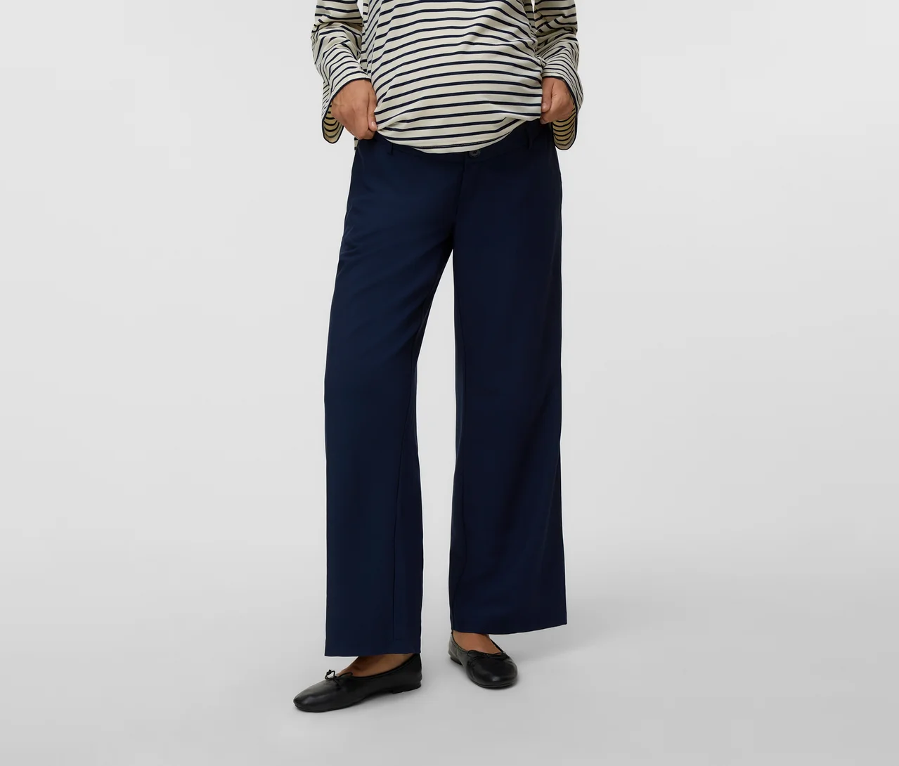 Personne portant un haut rayé à manches longues, un pantalon de maternité large bleu marine et des ballerines noires, ajustant la taille du pantalon.