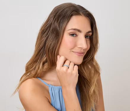 Une femme aux cheveux ondulés et au débardeur bleu touche son doigt orné d'une bague devant un fond clair.