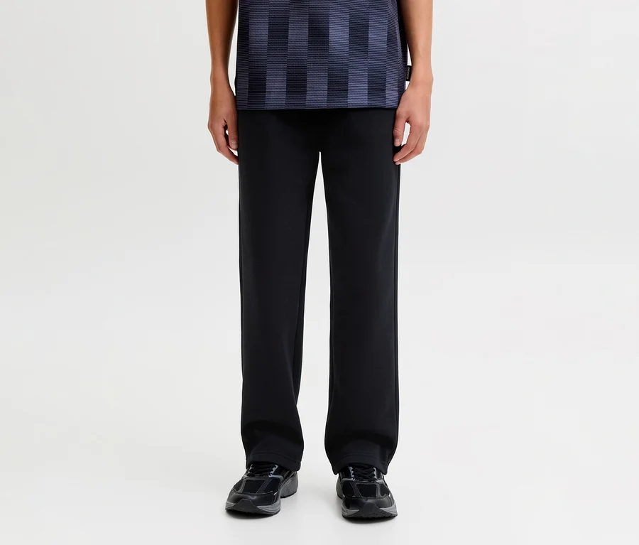 Personne debout, portant un pantalon de survêtement noir Jack&Jones Bradley, un t-shirt bleu foncé à motifs et des baskets sombres.