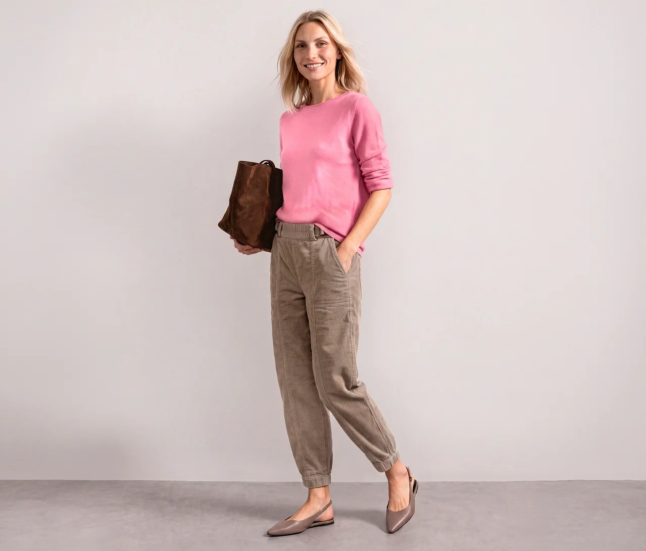 Femme souriante portant un pull en maille Street One rose pâle, un pantalon marron clair et des ballerines taupe, tenant un sac marron.