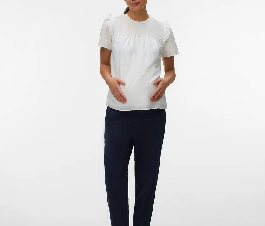 Femme enceinte debout, les mains sur le ventre, portant un haut blanc MAMALICIOUS « MLDORA » à manches courtes volantées et un pantalon bleu marine.