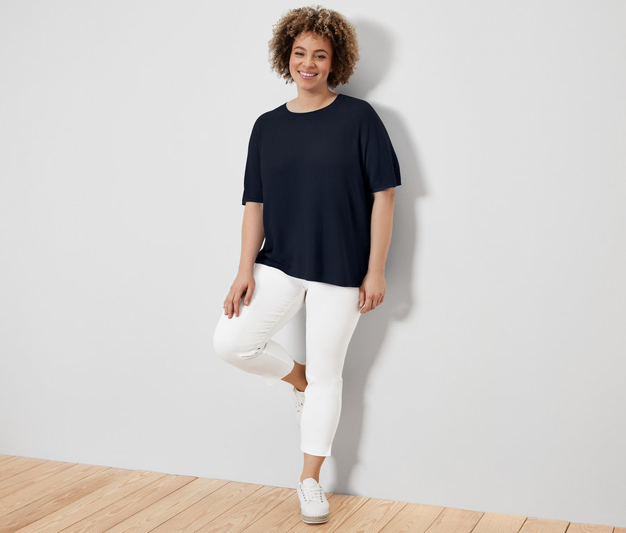 Une femme adossée à un mur porte un t-shirt en maille fine bleu marine, un pantalon blanc et des baskets.