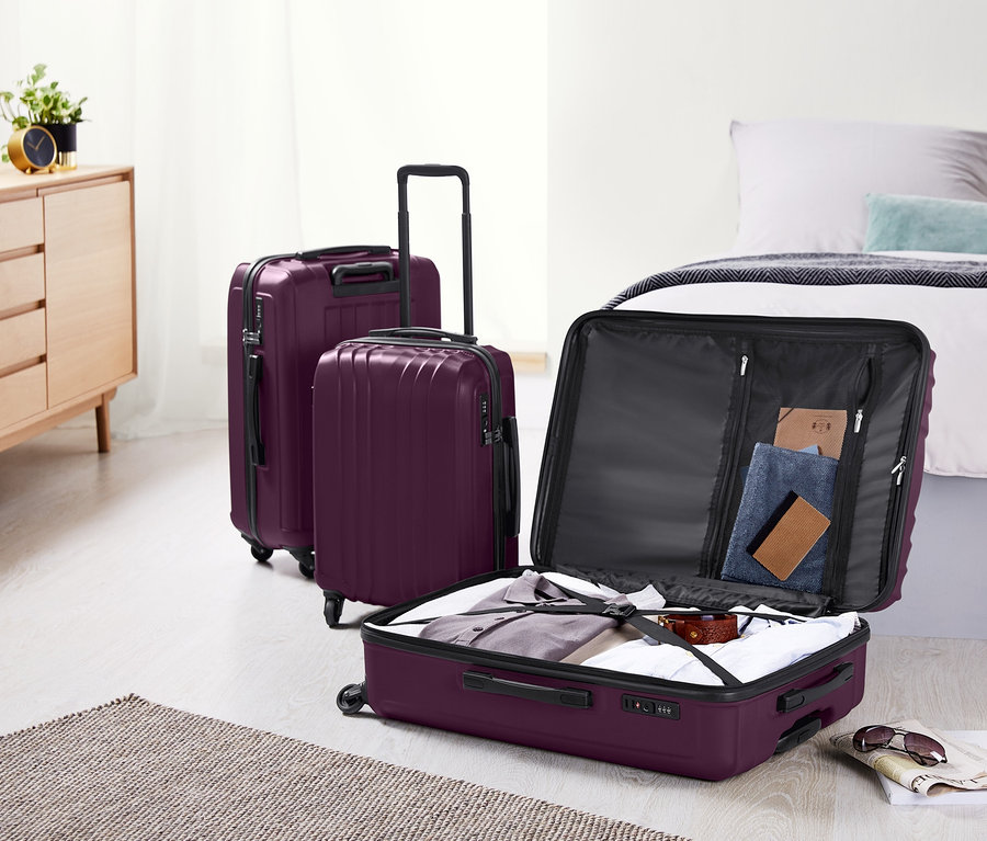 Trois valises rigides violettes (petite, moyenne et grande) sont posées dans une chambre à côté d'un lit et d'une commode. Une valise est ouverte et remplie de vêtements.