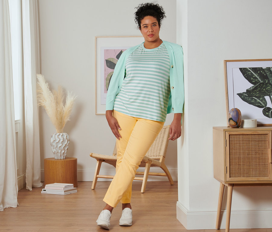 Une femme appuyée contre un mur porte un t-shirt à rayures vert menthe, un cardigan à encolure en V, un jean slim jaune et des baskets en cuir velours.