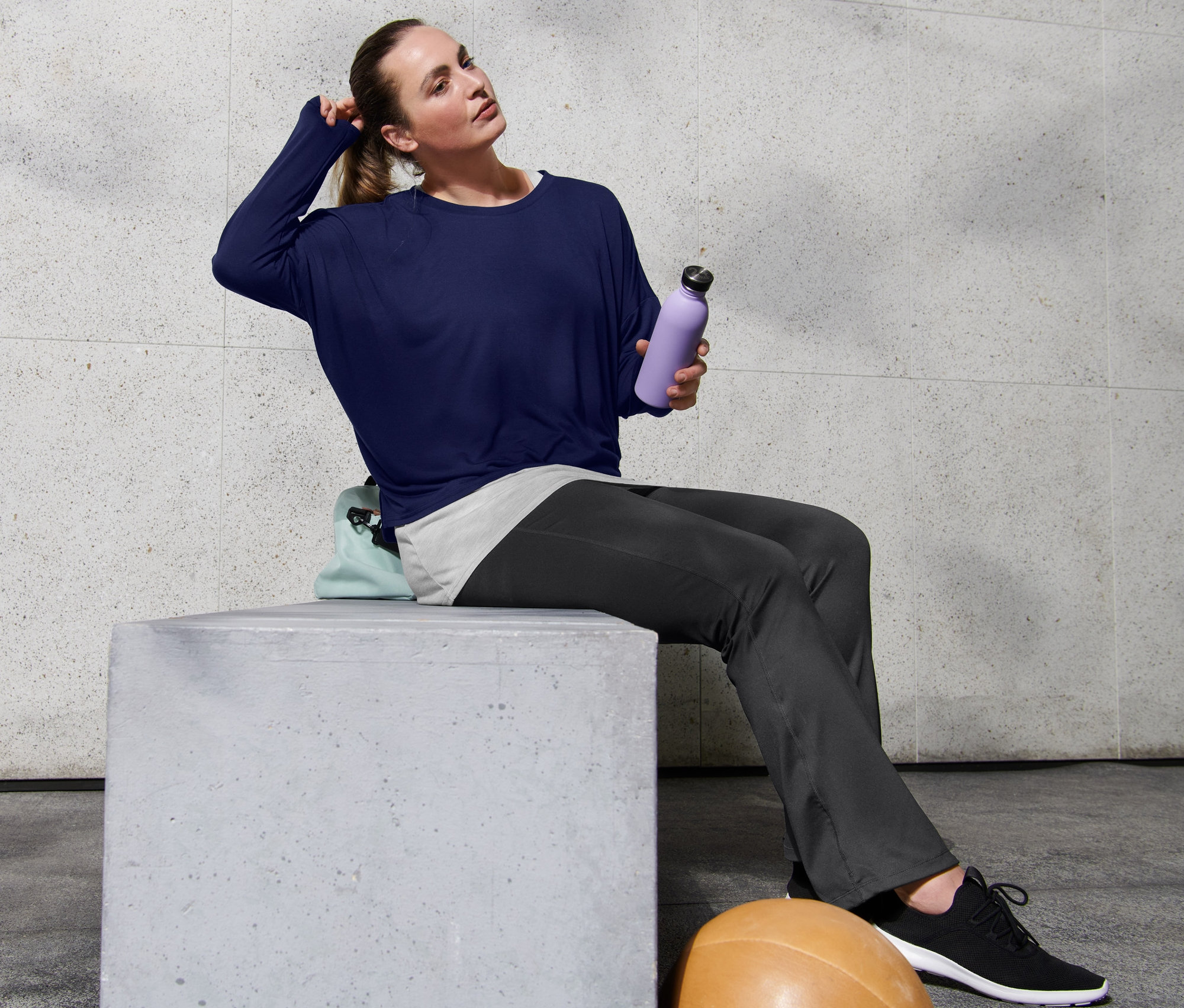 Femme assise sur un bloc de béton, portant un pantalon jazz de sport et un t-shirt de sport 2 en 1, tenant une bouteille d'eau violette à la main.