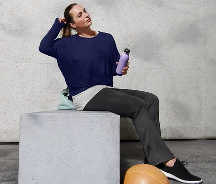 Femme assise sur un bloc de béton, portant un pantalon jazz de sport et un t-shirt de sport 2 en 1, tenant une bouteille d'eau violette à la main.