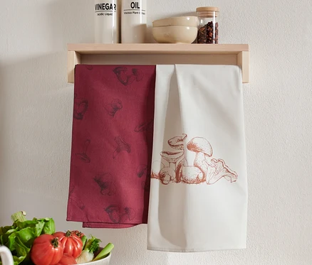 Deux linges de cuisine suspendus à une étagère en bois. L'un est bordeaux avec un imprimé de champignons, l'autre est blanc avec une illustration de champignons.