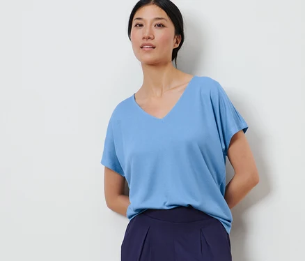 Une femme pose sur un fond blanc, portant une blouse structurée bleu clair et un pantalon bleu marine.