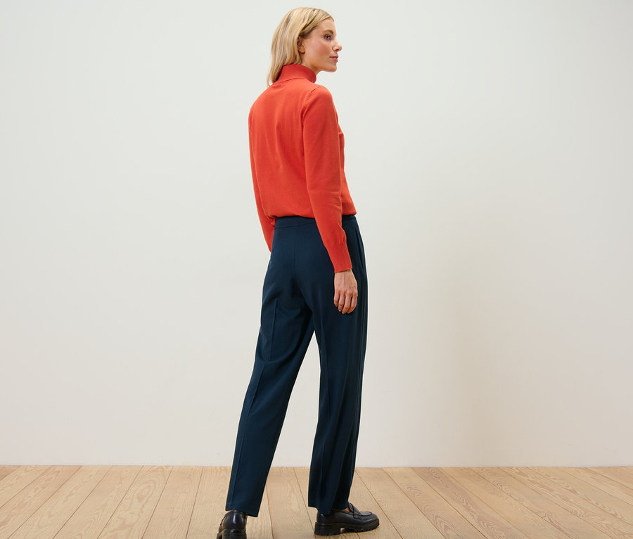 Une femme se tient dos à la caméra portant un pull à col roulé orange et un pantalon en jersey marine avec passepoil.