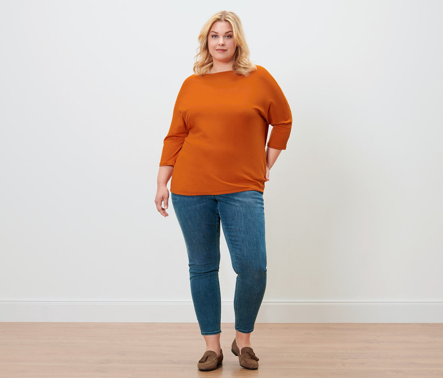 Une femme pose avec un pull en maille fine orange, un tregging « Fit Nelly » et des chaussures en cuir velours.