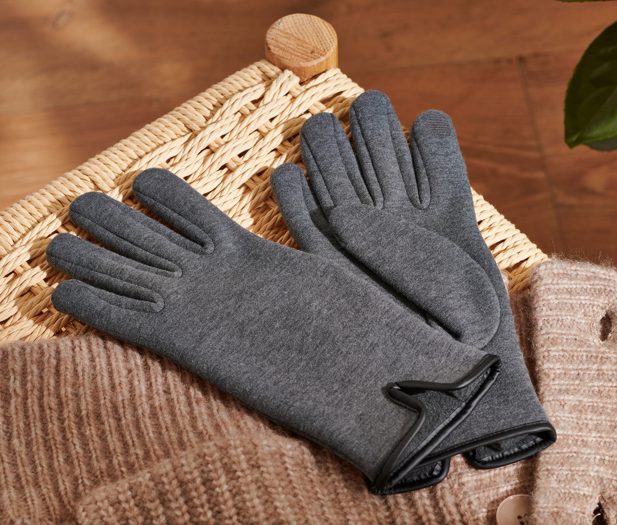 Des gants sweat anthracite reposent sur un panier en osier à côté d'un pull beige.