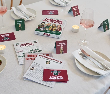 Sur une table se trouvent le jeu d'enquête criminelle « Mordspektakel » (en allemand), des cartes de personnages, des marque-places et des assiettes avec des couverts.