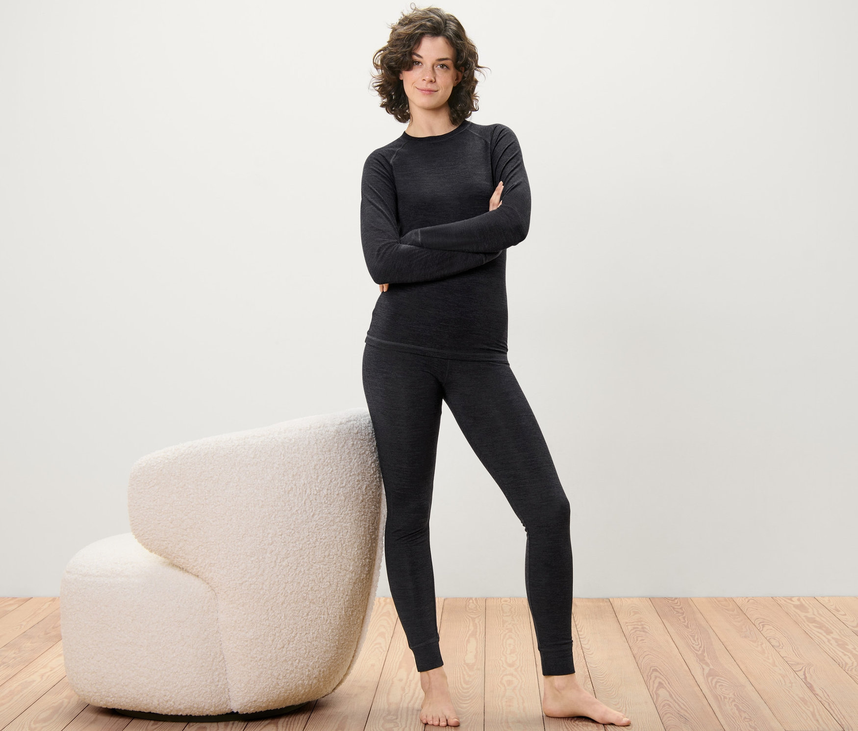 Une femme pose en caleçon long en laine certifiée RWS et haut sur un plancher en bois à côté d'un fauteuil crème.