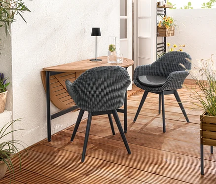 Deux chaises à coque avec tressage en polyrotin sont placées près d'une table repliable «
AElin
A». Une lampe de table d’extérieur à LED rechargeable noire est posée sur la table. Un bac à plantes «
AElin
A» est au premier plan.