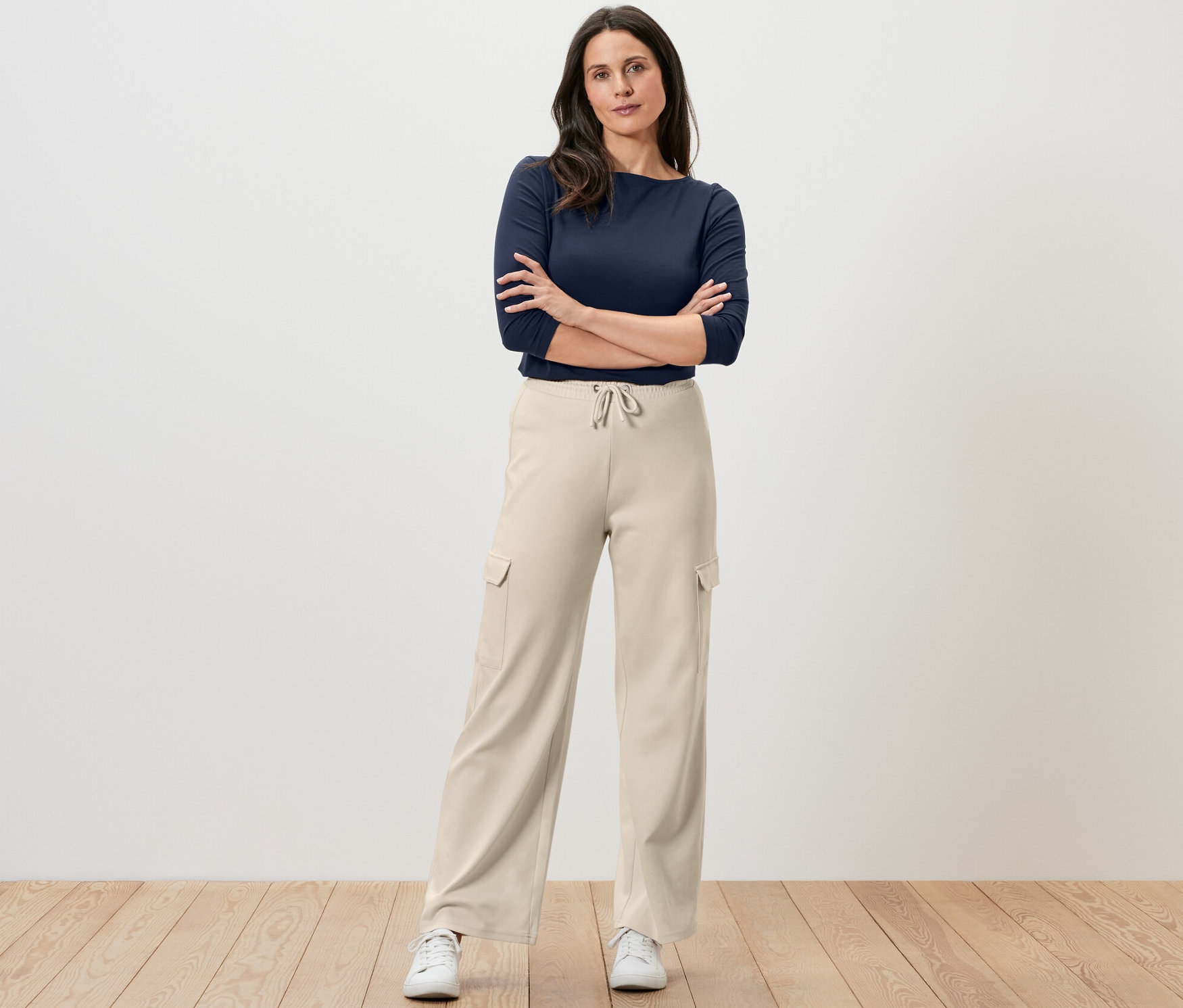 Une femme se tient debout, les bras croisés, portant un pantalon molletonné blanc crème avec des poches cargo et un haut bleu marine.