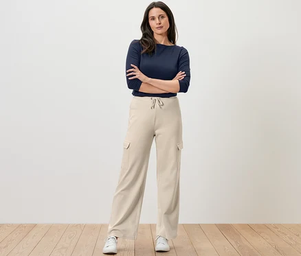 Une femme se tient debout, les bras croisés, portant un pantalon molletonné blanc crème avec des poches cargo et un haut bleu marine.