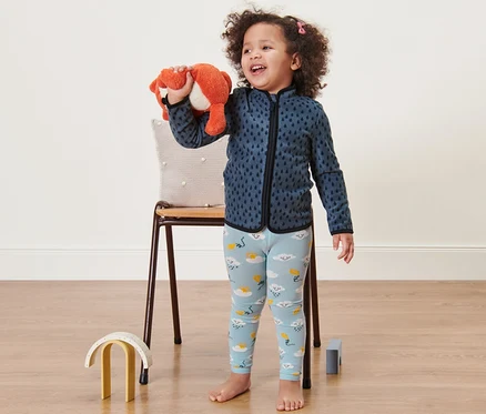 Petite fille souriante tenant un renard en peluche. Elle porte des leggings bleus avec des nuages et un sweat-shirt bleu.
