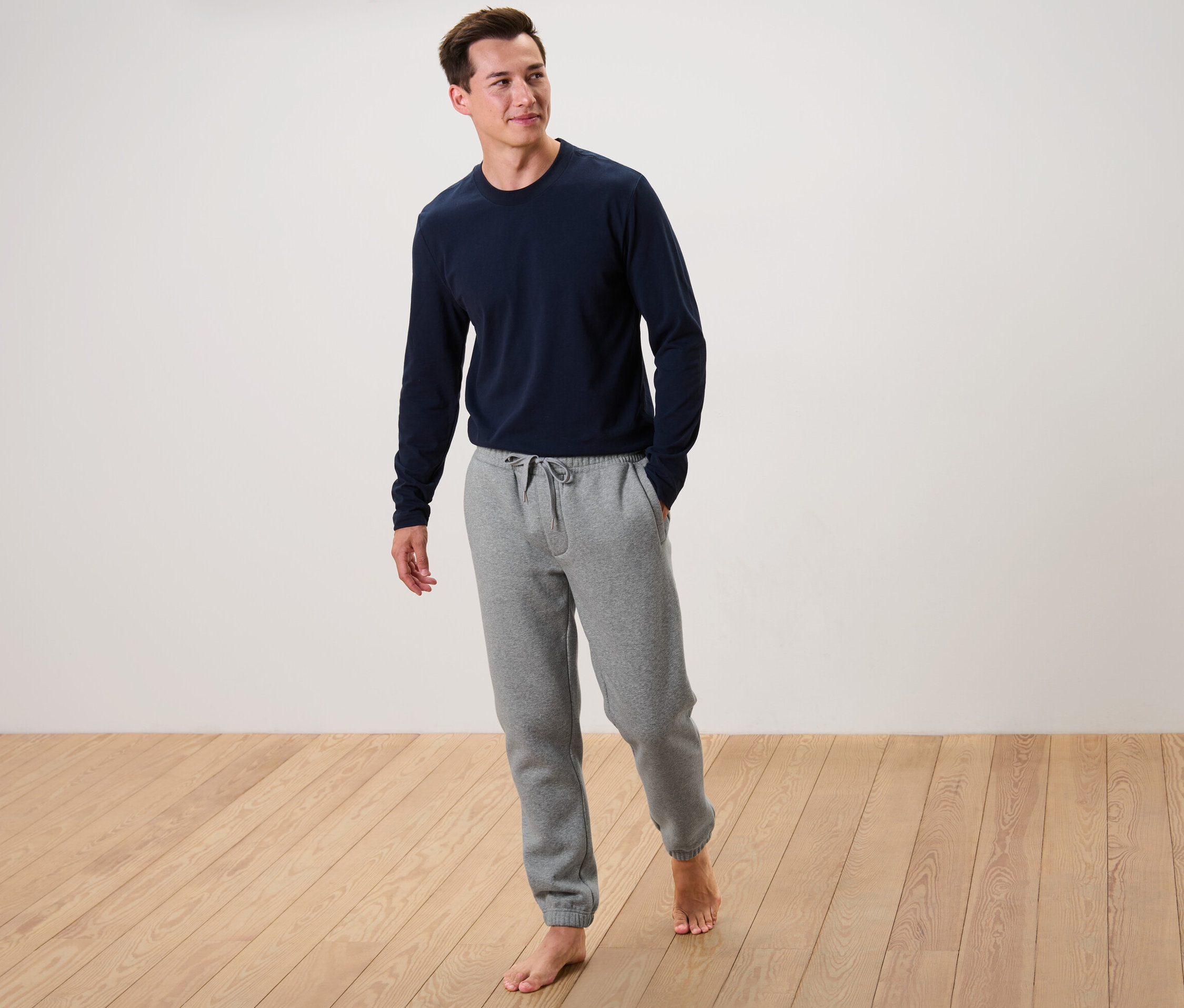 Un homme se tient pieds nus sur un plancher en bois, portant un t-shirt bleu à manches longues et un pantalon de survêtement gris.