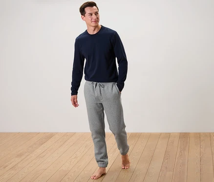 Un homme se tient pieds nus sur un plancher en bois, portant un t-shirt bleu à manches longues et un pantalon de survêtement gris.