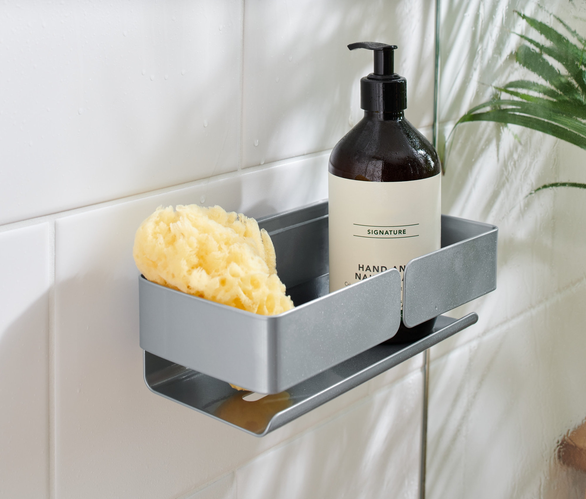 Une tablette de douche grise fixée sur des carreaux blancs contient un flacon brun à pompe et une éponge jaune.