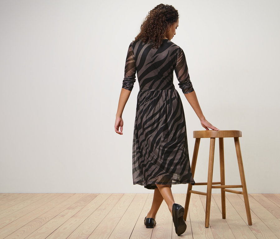 Femme posant dans une robe en mesh imprimée, une main reposant sur un tabouret en bois.
