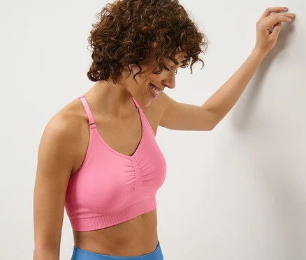 Une femme souriante, appuyée contre un mur, porte une brassière de sport Seamless rose vif.