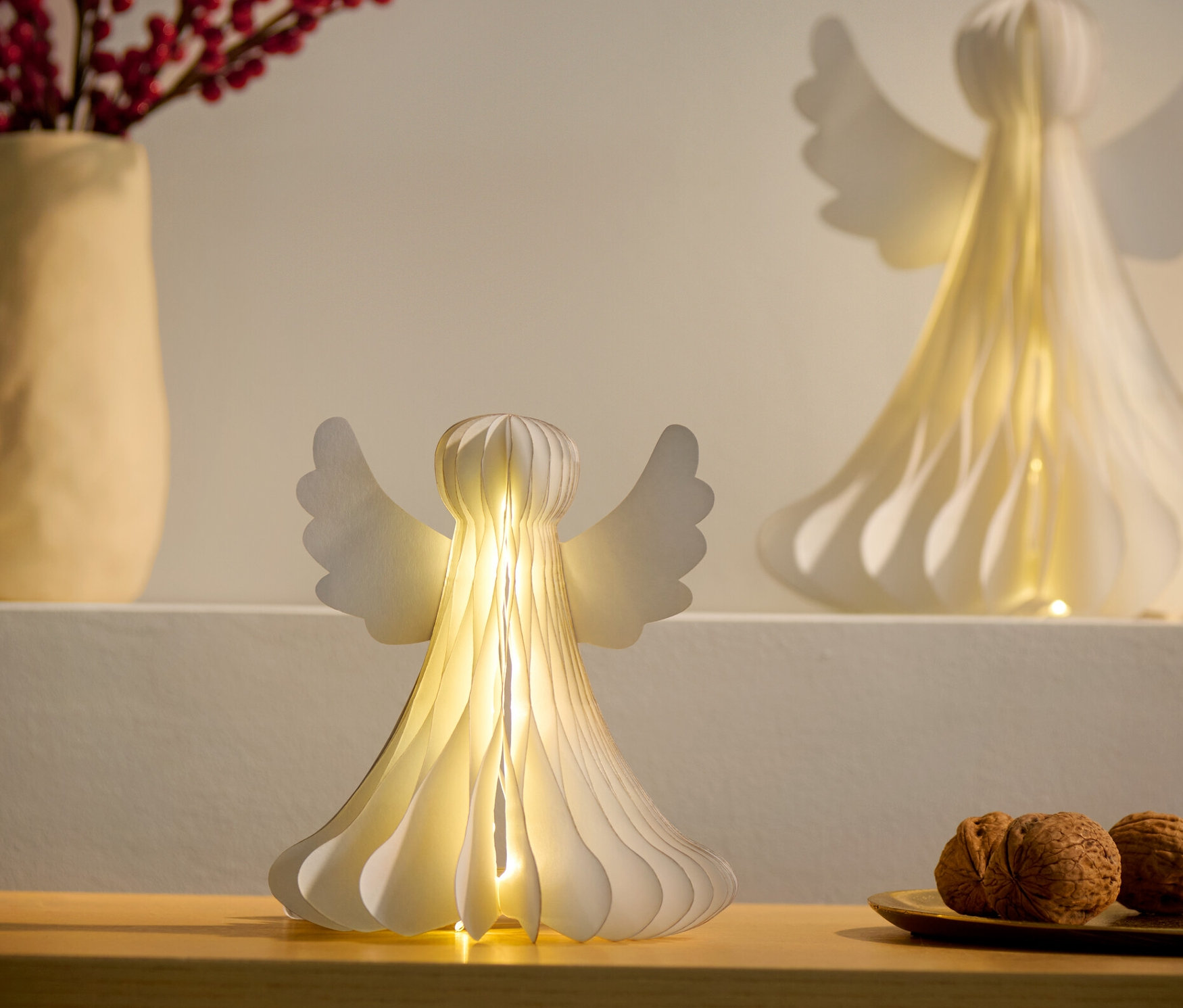 Deux petits anges en papier LED, l'un sur une armoire et l'autre sur une étagère, à côté d'un vase avec des baies rouges et d'une assiette de noix.