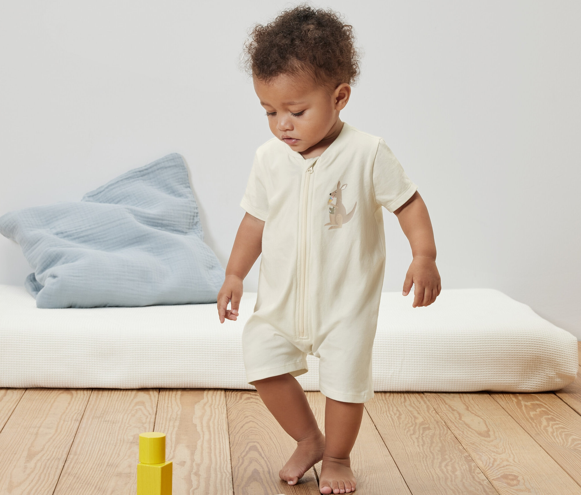 Un bébé portant un pyjama court pour bébé avec un imprimé kangourou, debout sur un plancher de bois près de blocs de bois.