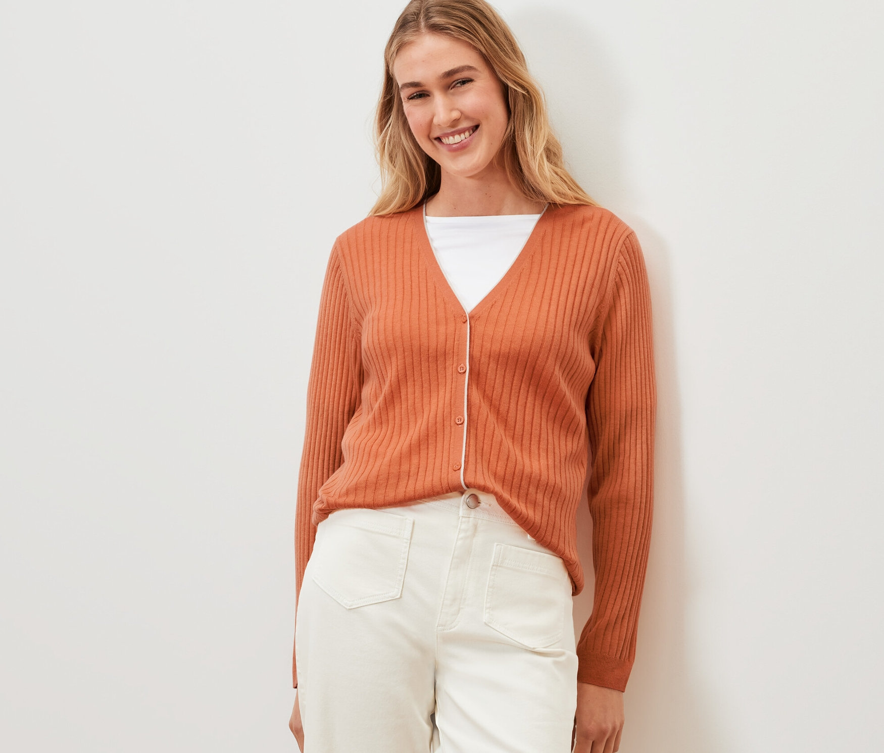 Une femme pose en cardigan en soie orange et pantalon blanc.