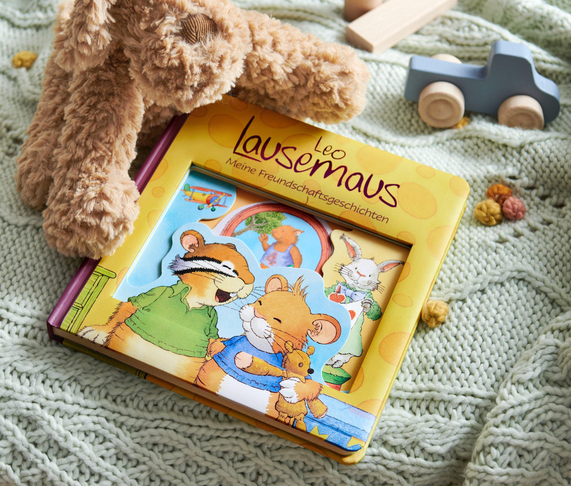 Un livre « Leo Lausemaus – meine Freundschaftsgeschichten » est posé sur une couverture tricotée avec un ours en peluche et une voiture en bois.