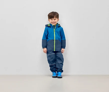 Garçon souriant posant en veste bleue à capuche et pantalon bleu marine avec bandes réfléchissantes.