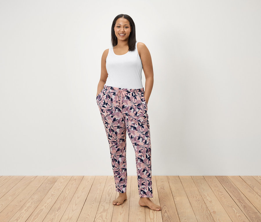 Une femme se tient debout, les mains dans les poches d'un pantalon à motifs floraux. Elle porte un débardeur blanc.