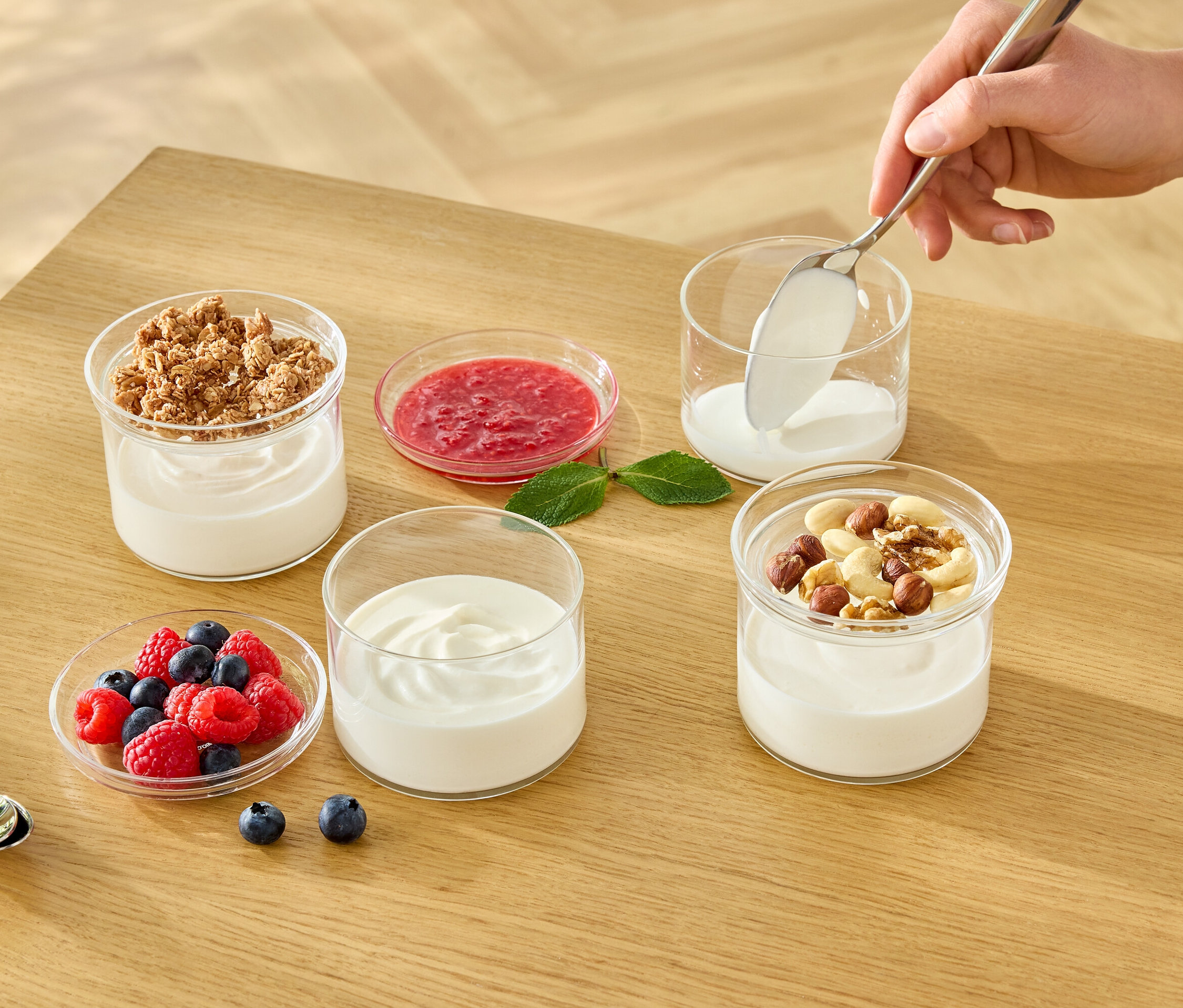Une main met du yaourt blanc dans un verre avec une cuillère. Sur la table, il y a d'autres verres avec du yaourt, du muesli, des fruits et des noix.
