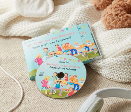 Un livre audio « Wir Kinder aus dem Möwenweg – Sommerzeit und Ferienspaß » et des écouteurs sont posés sur une couverture à côté d'une peluche.