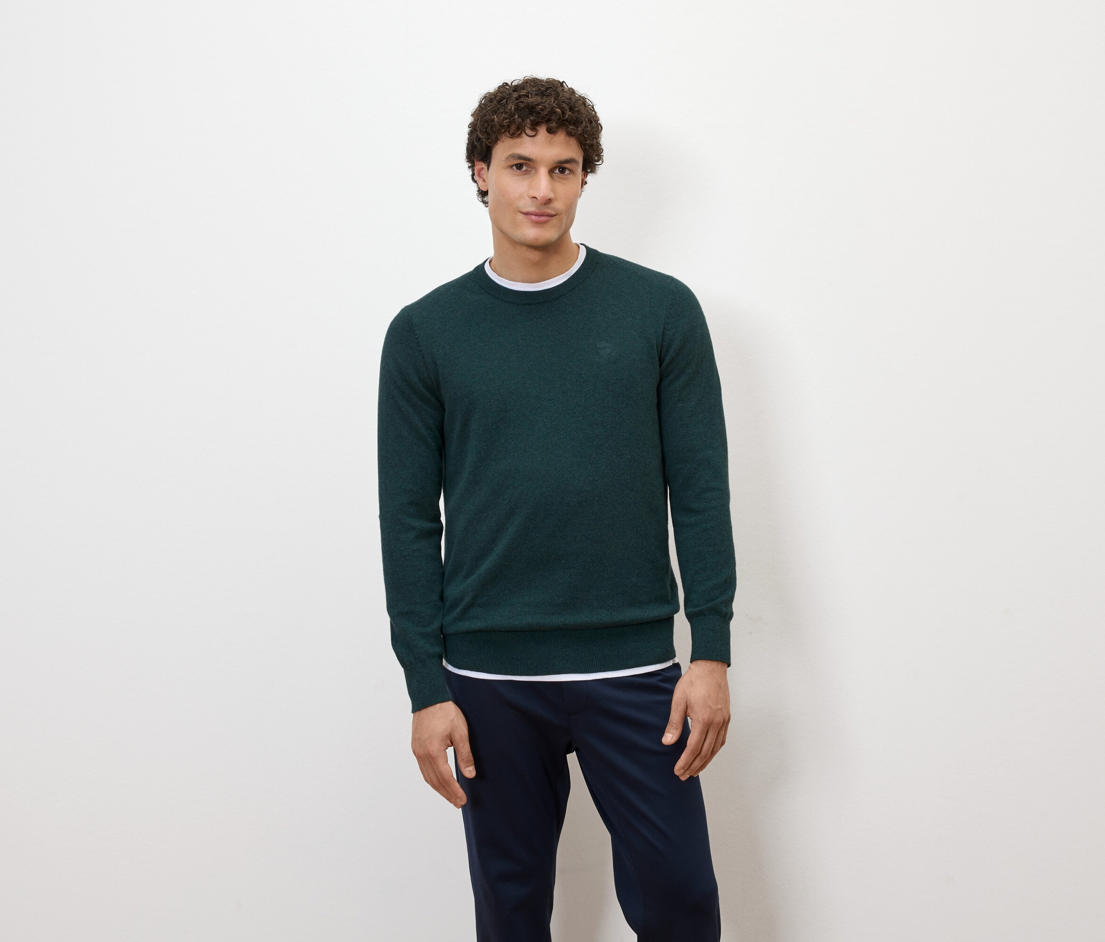 Un homme pose devant un fond blanc, portant un pull vert foncé et un pantalon bleu marine.