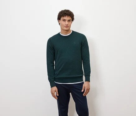 Un homme pose devant un fond blanc, portant un pull vert foncé et un pantalon bleu marine.