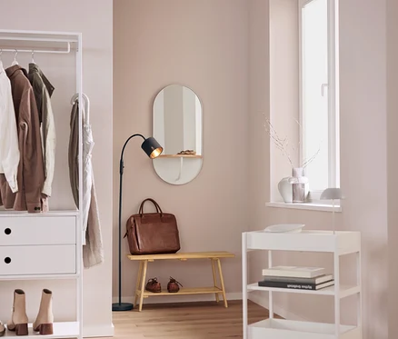 Dans un intérieur, on trouve une étagère blanche avec des vêtements, un miroir ovale, un sac en cuir marron sur un banc, une lampe noire et un chariot à roulettes blanc.