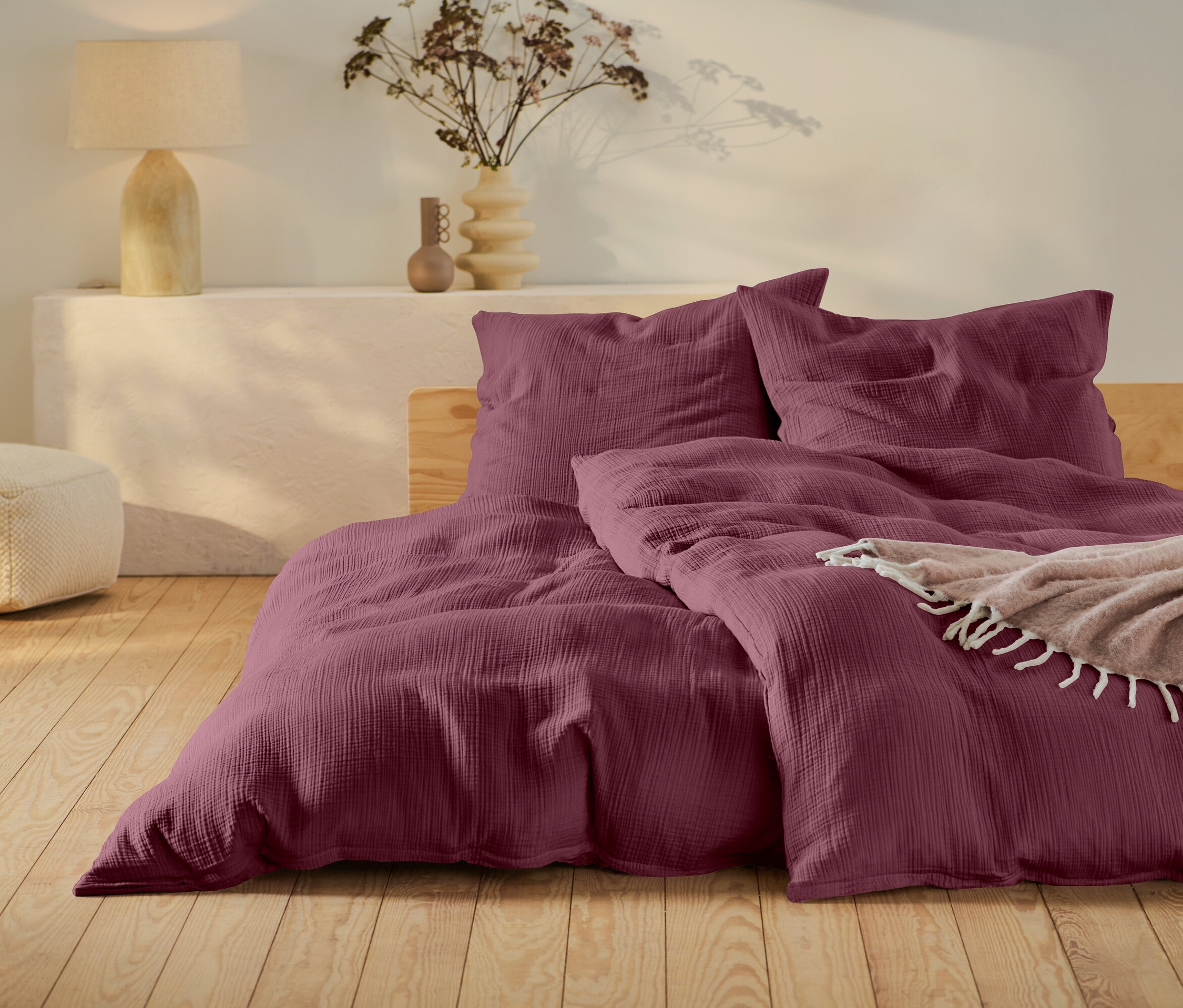 Linge de lit bordeaux sur un lit avec cadre en bois, un pouf beige et une lampe sur une table blanche.