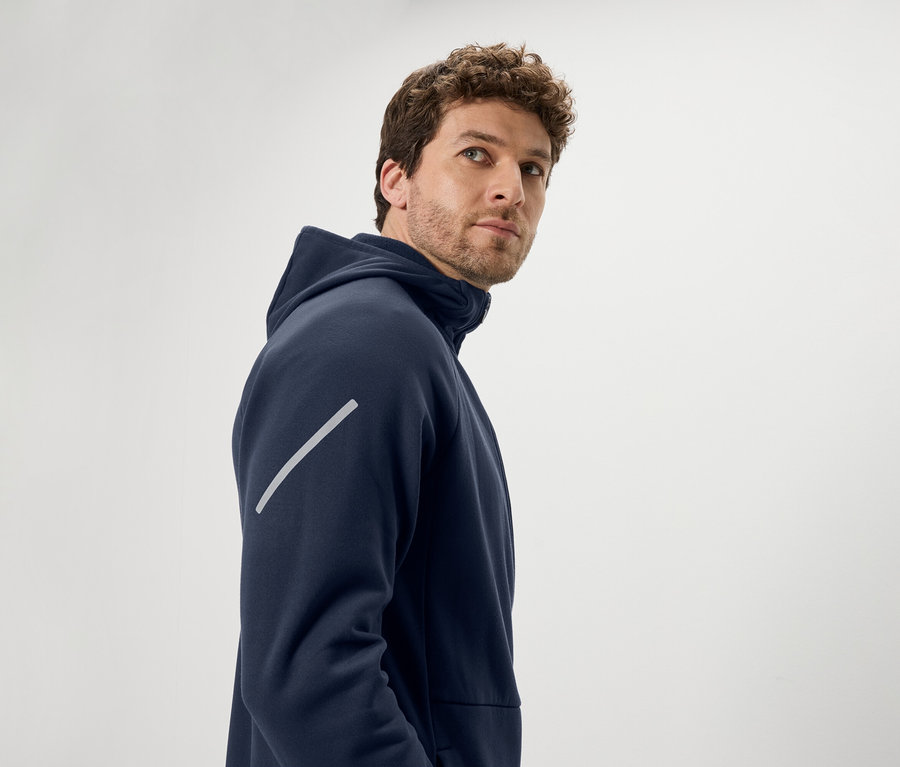 Un homme aux cheveux bouclés bruns regarde par-dessus son épaule. Il porte un sweat à capuche bleu marine avec une bande réfléchissante diagonale sur le bras.