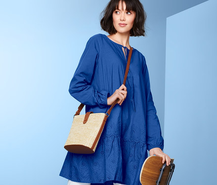 Femme posant sur une chaise en chemisier long à broderie bleu et pantalon blanc.