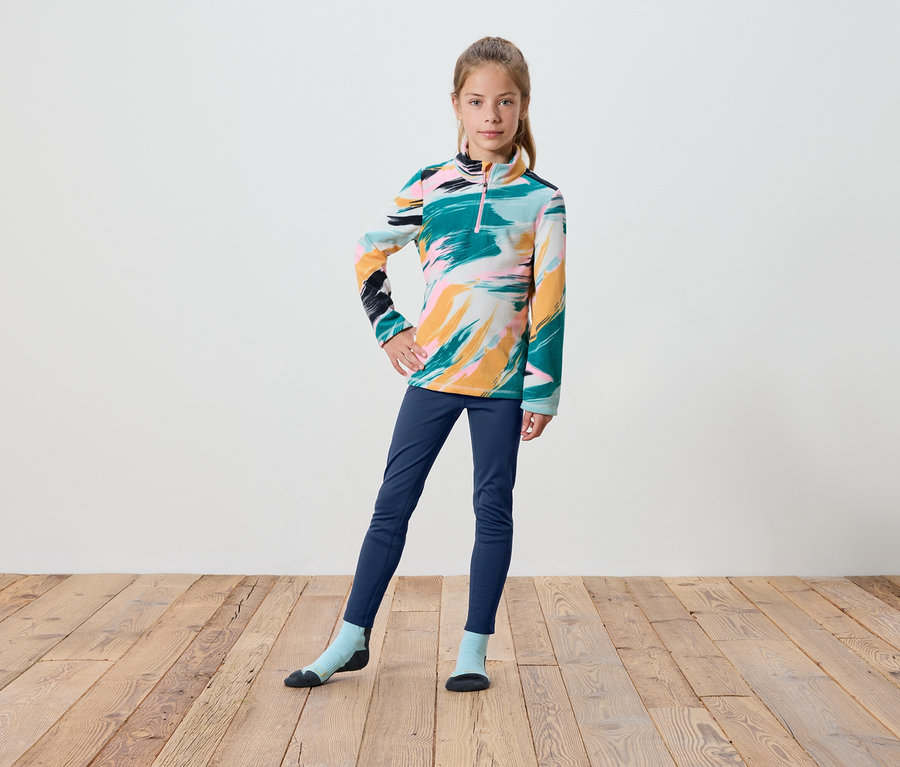 Une fille portant un pull coloré à manches longues, un pantalon bleu marine et des chaussettes se tient sur un plancher en bois.