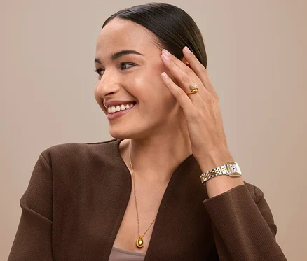 Femme souriante touchant son visage portant une bague, une montre et un collier en or.