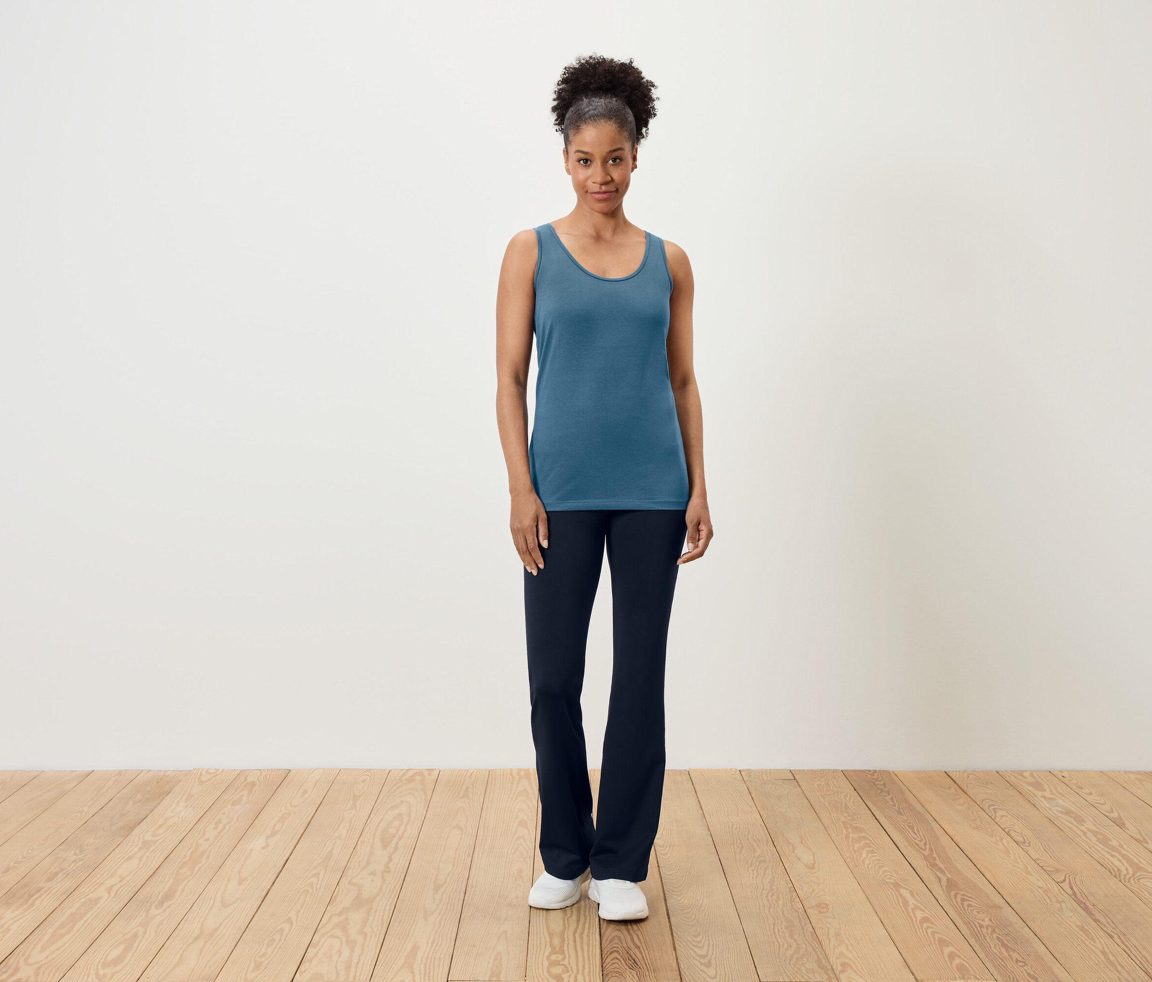 Une femme se tient sur un plancher en bois, portant un débardeur bleu et un pantalon bleu marine.