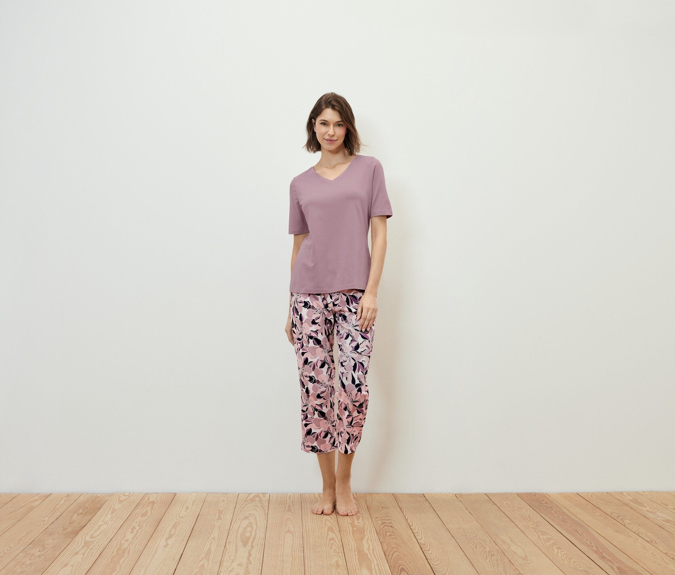 Une femme debout porte un t-shirt violet et un pantalon à fleurs.