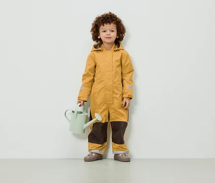 Un enfant aux cheveux bouclés se tient debout dans une combinaison jaune et tient un arrosoir vert clair.